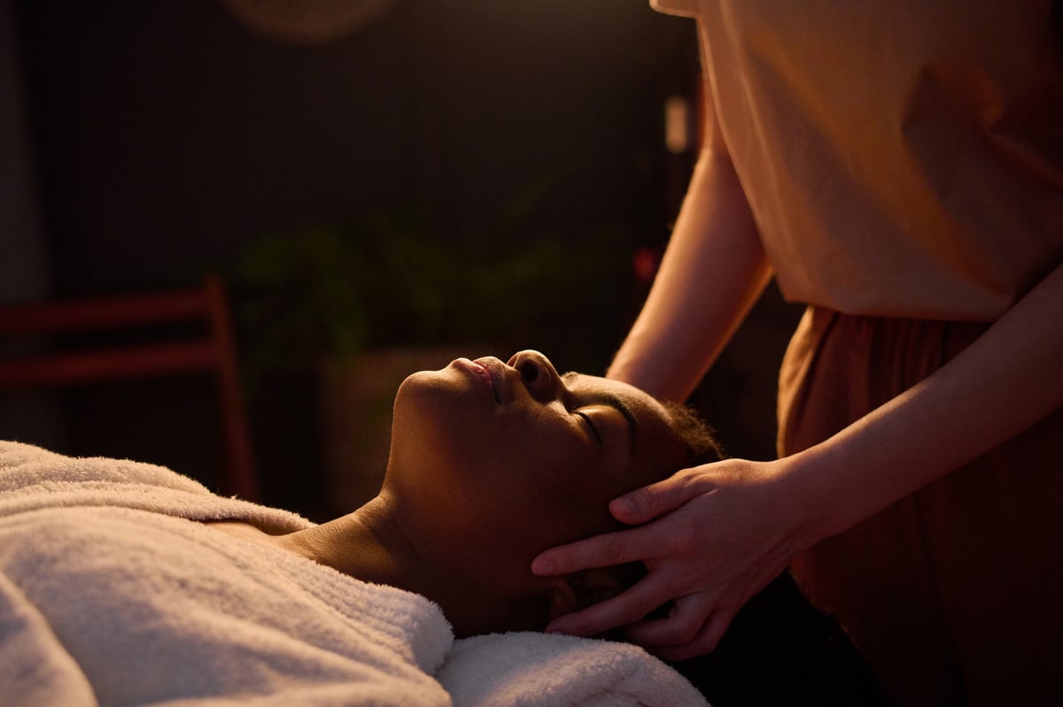 African American woman in bathrobe lying with closed eyes enjoying head massage by professional masseuse